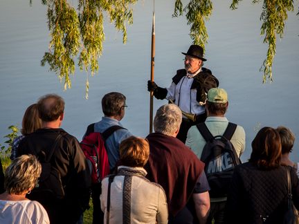 Ein Mensch in alter Kleidung mit Schwert spricht vor einer Gruppe - eine Stadtführung. Die Gruppe steht am Ufer eines ruhigen Sees.