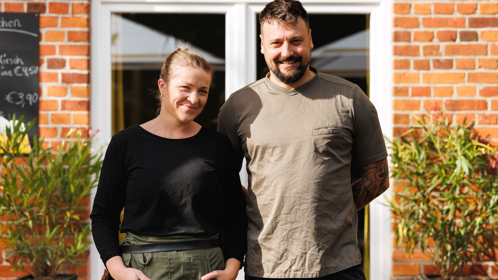 Jusitne und Felix Duwe_landhaus Köthel Ein Mann und eine Frau vor einer Backsteinmauer mit Eingangstür.