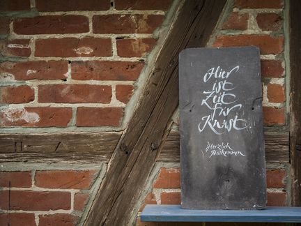 Eine alte Schiefertafel hängt an einer Backsteinwand. Darauf steht: ‘Hier ist Zeit für Kunst’. Unten steht kleiner: ‘Herzlich Willkommen’.