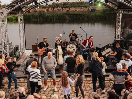Viele Menschen tanzen fröhlich vor einer Bühne. Auf der Bühne spielt eine Band. Es ist eine offene Veranstaltung.