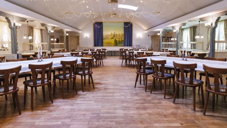 Ein großer, heller Saal mit vielen Tischen und Stühlen. Die Tische sind mit weißen Decken und Kerzen für Gäste vorbereitet. Vorne ist eine Bühne mit einem großen Bild von einer Landschaft.