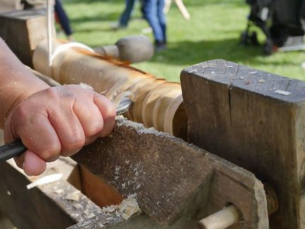 „Eine Hand arbeitet mit einem Werkzeug an einem Stück Holz. Im Hintergrund stehen Menschen auf einer Wiese.